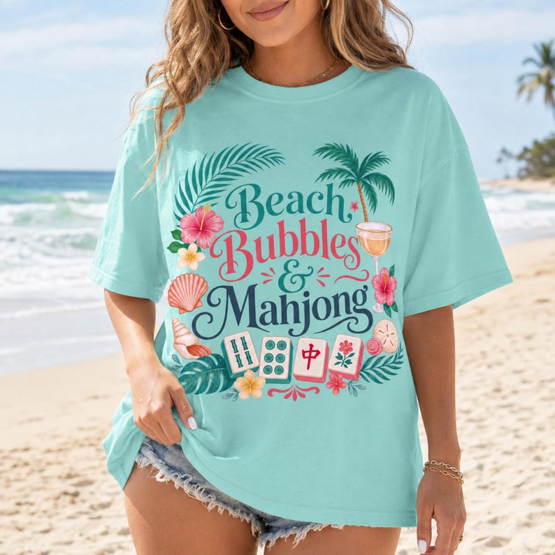 May include: A turquoise t-shirt featuring the text "Beach Bubbles & Mahjong" surrounded by tropical illustrations. The design includes palm trees, flowers, and seashells. The shirt has a loose fit, ideal for a day at the seaside.