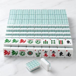 K&ouml;nnte beinhalten: Ein komplettes Mahjong-Set mit wei&szlig;en Spielsteinen mit roten, gr&uuml;nen und schwarzen Symbolen und Schriftzeichen. Das Set enth&auml;lt einen Stapel passender teal- und wei&szlig; karierter Spielsteinhalter. Die Spielsteine sind auf einer wei&szlig;en Oberfl&auml;che angeordnet.