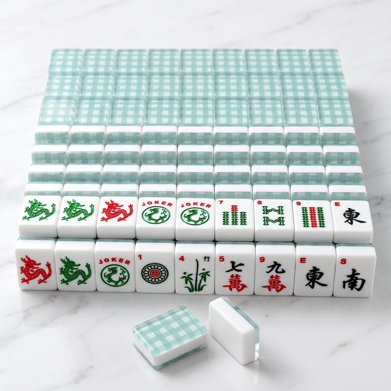 K&ouml;nnte beinhalten: Ein komplettes Mahjong-Set mit wei&szlig;en Spielsteinen mit roten, gr&uuml;nen und schwarzen Symbolen und Schriftzeichen. Das Set enth&auml;lt einen Stapel passender teal- und wei&szlig; karierter Spielsteinhalter. Die Spielsteine sind auf einer wei&szlig;en Oberfl&auml;che angeordnet.