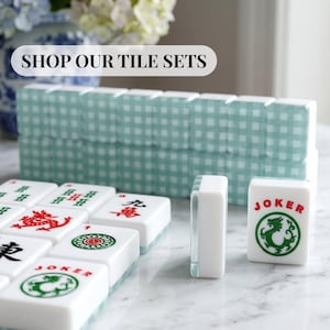 May include: A collection of white Mahjong tiles with red and green symbols and characters. The image includes a stack of tiles with a light blue gingham pattern and the text "SHOP OUR TILE SETS".