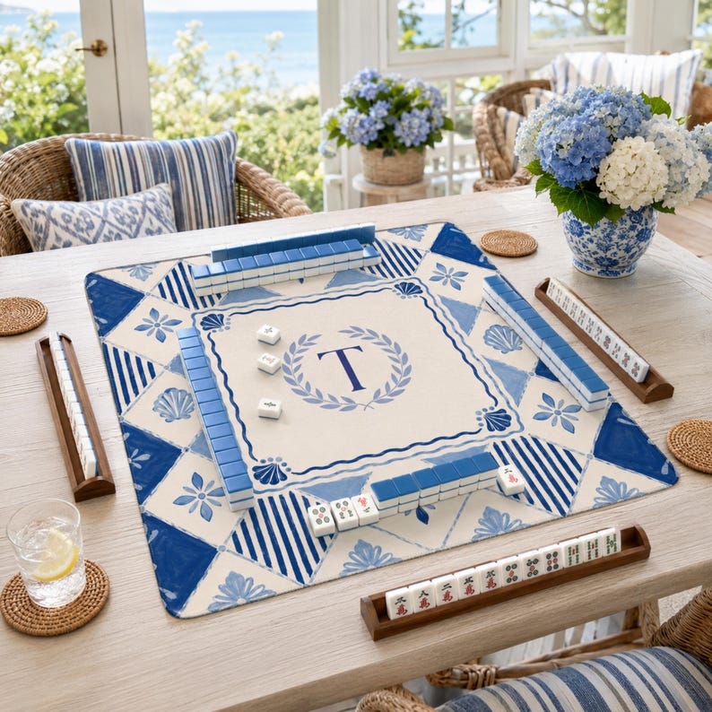 Op de afbeelding: Een Mahjong-spelset op een lichte houten tafel. Het spelbord heeft een blauw en wit geometrisch ontwerp met een cr&egrave;mekleurig centraal vierkant met de letter "T" in een krans. Blauwe tegels zijn rond het bord gerangschikt, met houten tegelhouders.