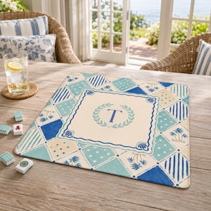 May include: A square placemat with a blue and cream geometric pattern. The center features a wreath design with the letter "T". The placemat has a coastal theme with palm trees and seashell accents.