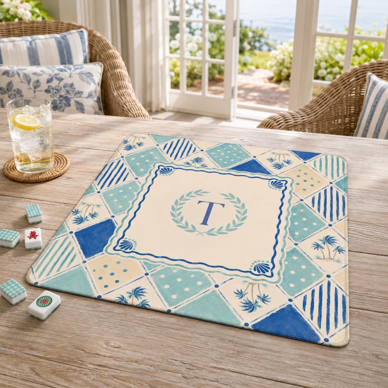 May include: A square placemat with a blue and cream geometric pattern. The center features a wreath design with the letter "T". The placemat has a coastal theme with palm trees and seashell accents.