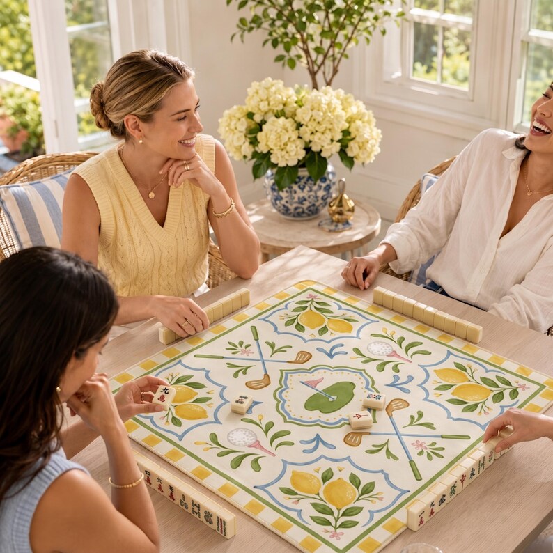 May include: A square mahjong game board with a golf theme, featuring lemons and golf clubs. The board is surrounded by players. The game is set on a light-colored wooden table. The board has a green, yellow, and white color scheme.