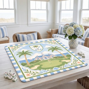 May include: A square golf-themed placemat with a blue and white checkered border. The center features a golf course scene with palm trees, a clubhouse, and a golf flag. A golf club and palm tree crest is at the top. A few game tiles are on the table.