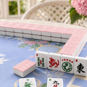 May include: A Mahjong set with white tiles and pink patterned backs is displayed. Some tiles are arranged in rows, while others are scattered. The tiles feature various symbols and characters, including dragons and the word "Joker".