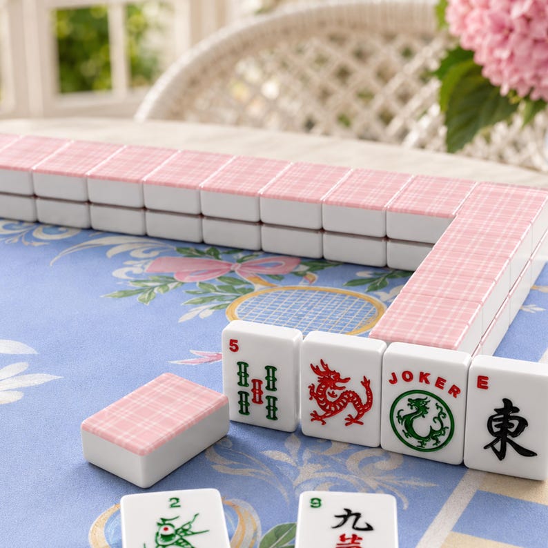 May include: A Mahjong set with white tiles and pink patterned backs is displayed. Some tiles are arranged in rows, while others are scattered. The tiles feature various symbols and characters, including dragons and the word "Joker".