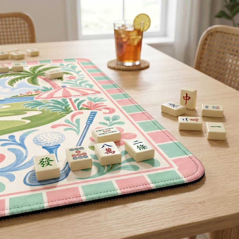 May include: A colorful Mahjong game set on a table. The game mat features a golf course design with a pink and green border. Several ivory-colored tiles are scattered around the mat, and a glass of iced tea sits nearby.