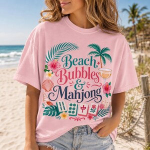 May include: Pale pink t-shirt featuring the text "Beach Bubbles & Mahjong" in a decorative font, with tropical illustrations. The design includes palm trees, flowers, seashells, and mahjong tiles. The shirt is worn by a person on a beach.