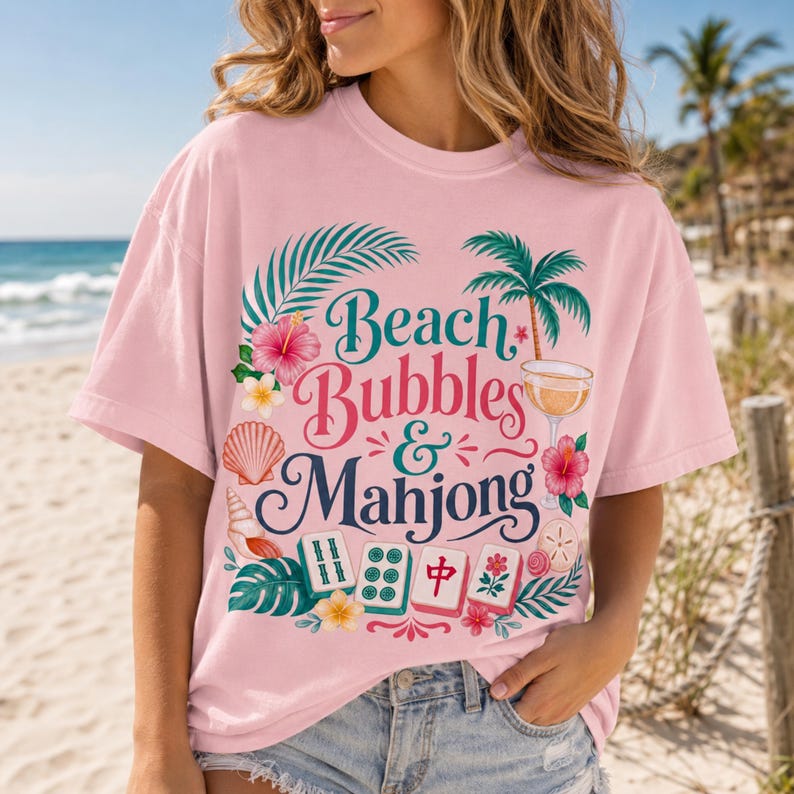 May include: Pale pink t-shirt featuring the text "Beach Bubbles & Mahjong" in a decorative font, with tropical illustrations. The design includes palm trees, flowers, seashells, and mahjong tiles. The shirt is worn by a person on a beach.