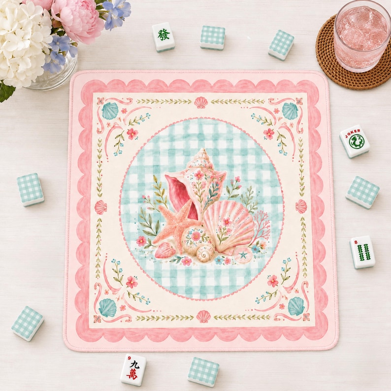 May include: A pink and cream-colored square mat with a scalloped edge, featuring a watercolor design of seashells, starfish, and floral elements. The center has a blue and white checkered pattern. Mahjong tiles are scattered around the mat.
