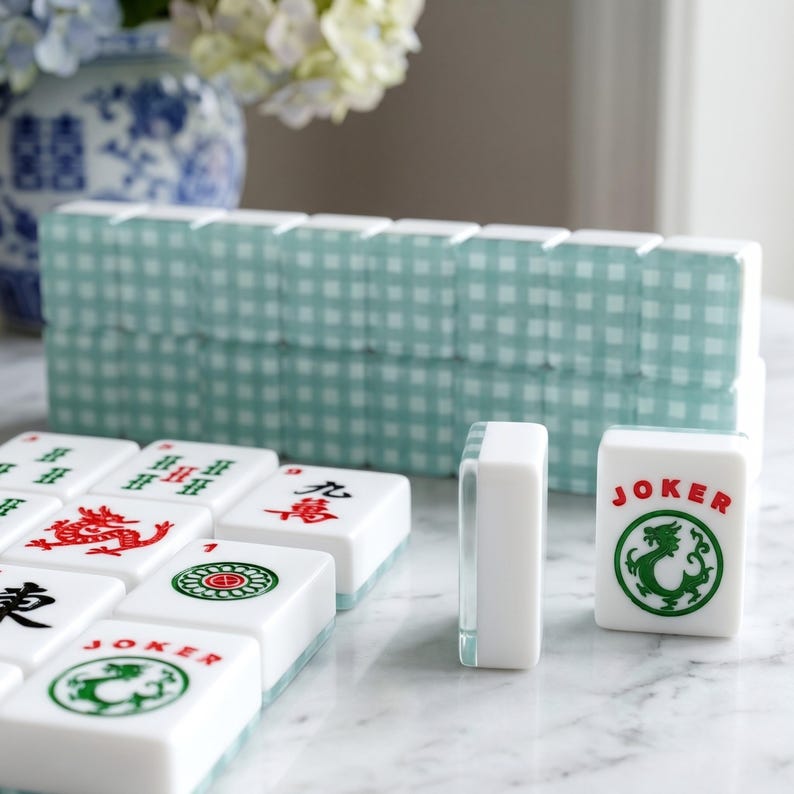 K&ouml;nnte beinhalten: Ein Satz wei&szlig;er Mahjong-Steine mit roten und gr&uuml;nen Symbolen und Zeichen. Die Steine sind auf einer wei&szlig;en Oberfl&auml;che angeordnet, mit einem Stapel hellblau-wei&szlig; karierter Steinehalter im Hintergrund. Ein Stein tr&auml;gt die Aufschrift "JOKER" mit einem gr&uuml;nen Drachen.