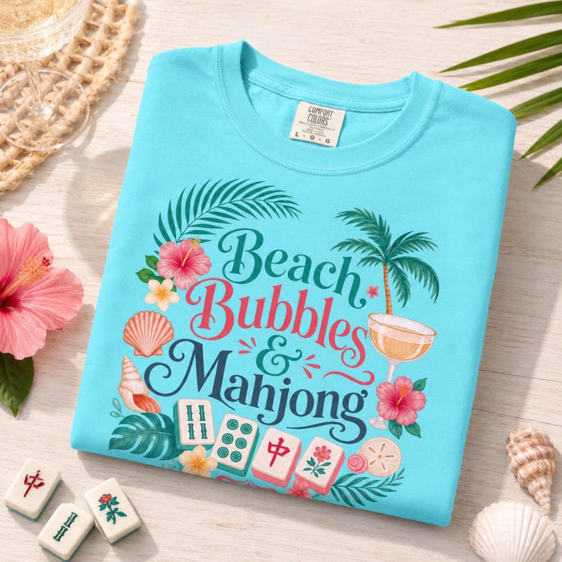 May include: A turquoise t-shirt featuring the text "Beach Bubbles & Mahjong" in a decorative font. The design includes palm trees, hibiscus flowers, seashells, and mahjong tiles. The shirt is made of a soft material.
