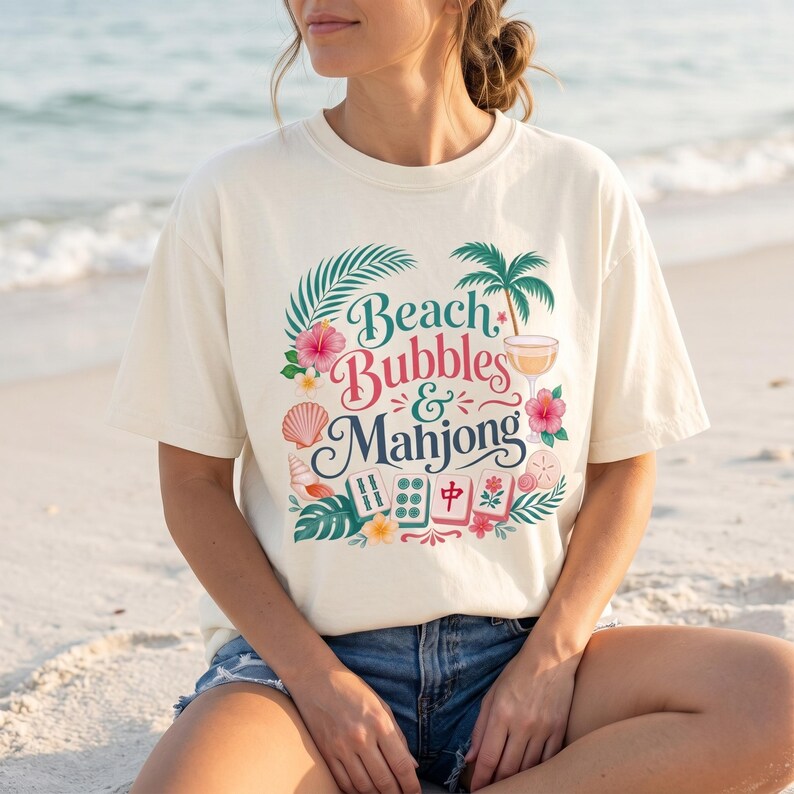 May include: A cream-coloured t-shirt with the text "Beach Bubbles & Mahjong" in a decorative design featuring tropical elements like palm trees, flowers, and seashells. The shirt is worn by a person sitting on a sandy beach.