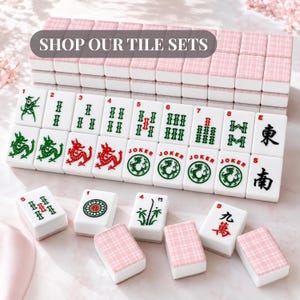 May include: A collection of white Mahjong tiles with various symbols and numbers, including dragons and Jokers. The tiles are arranged with pink and white patterned tile sets in the background. The image includes the text "SHOP OUR TILE SETS".