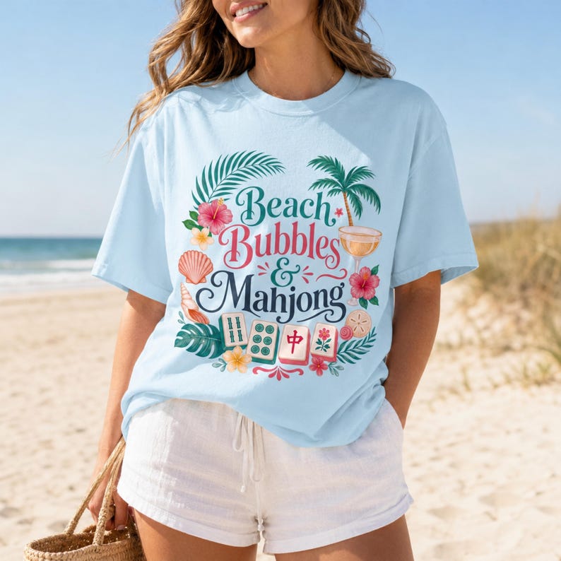 May include: Pale blue t-shirt featuring the text "Beach Bubbles & Mahjong" surrounded by tropical illustrations. The design includes a seashell, palm tree, and hibiscus flowers. The shirt is worn on a beach.