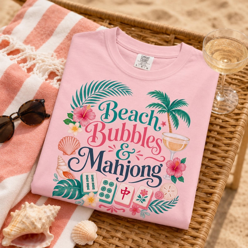 May include: A pale pink t-shirt with the text "Beach Bubbles & Mahjong" in a tropical design. The shirt features palm trees, flowers, and seashells. A glass of champagne and sunglasses are also visible.