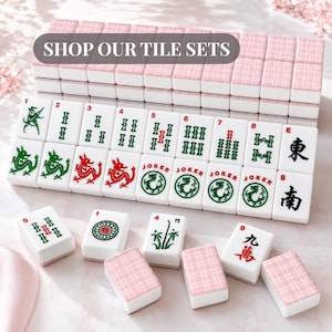 Pode incluir: Um conjunto de pe&ccedil;as de Mahjong brancas com v&aacute;rios s&iacute;mbolos e n&uacute;meros, incluindo drag&otilde;es e Jokers. As pe&ccedil;as s&atilde;o dispostas com conjuntos de pe&ccedil;as estampadas em rosa e branco no fundo. O texto "SHOP OUR TILE SETS" &eacute; exibido no topo.
