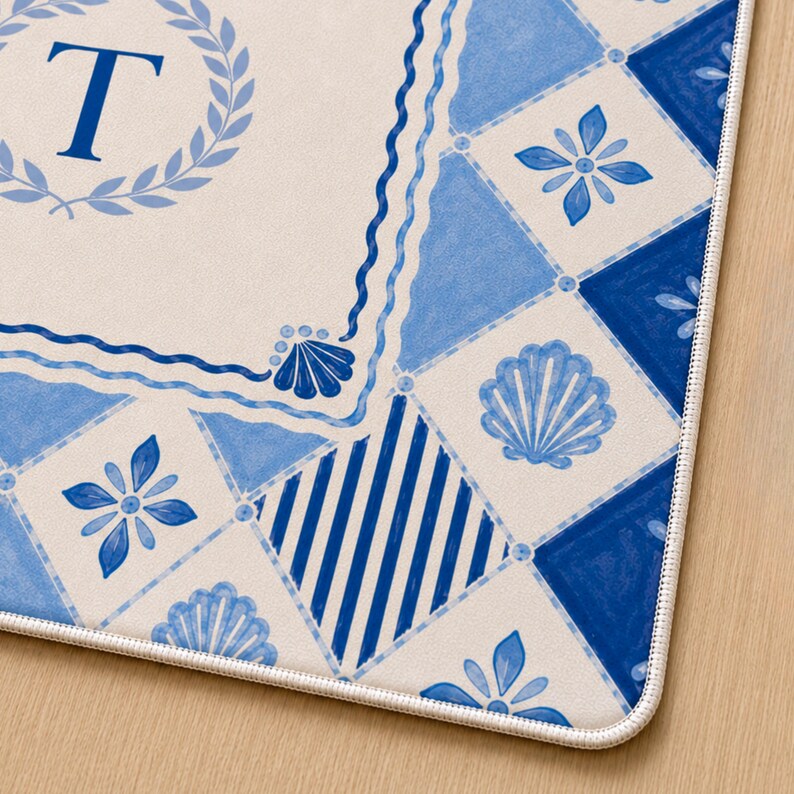 Op de afbeelding: Decoratieve mat met een blauw en wit nautisch ontwerp. De mat heeft een grote letter "T" in een lauwerkrans, samen met schelp- en bloemmotieven. De rand heeft een geschulpte rand.