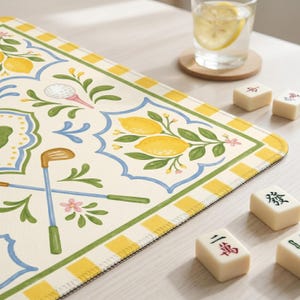 May include: A decorative table mat with a golf and lemon design. The mat features a cream background with illustrations of golf clubs, golf balls, and lemons. The border is yellow and green, with a blue decorative pattern. Mahjong tiles are scattered around the mat.