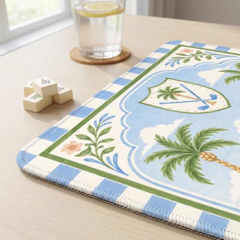 May include: A rectangular, decorative placemat with a blue and white checkered border. The center features a light blue background with palm trees, golf clubs, and floral accents. A glass of water with a lemon slice and mahjong tiles are also visible.