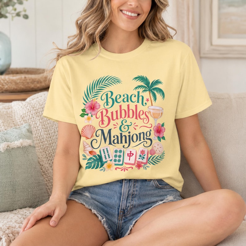 May include: Pale yellow t-shirt featuring the words "Beach Bubbles & Mahjong" in a tropical design. The design includes palm trees, flowers, seashells, and mahjong tiles. The shirt is a light yellow colour.