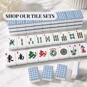 Op de afbeelding: Een complete Mahjong-tegelset met witte tegels met groene, rode en zwarte symbolen en karakters. De tegels zijn in rijen gerangschikt, sommige zijn opgestapeld. De set bevat tegels met de tekst "SHOP OUR TILE SETS".