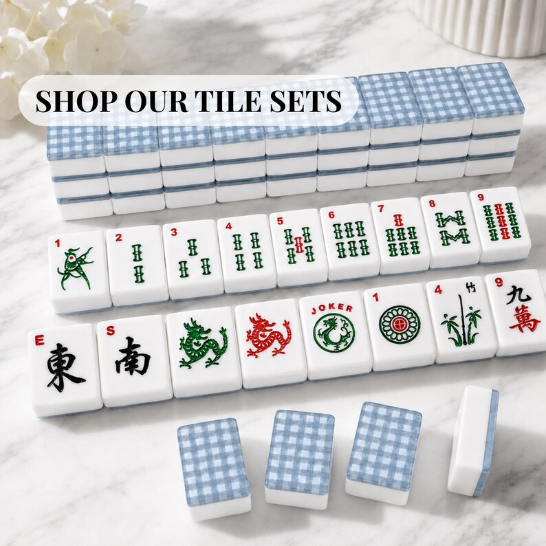 Op de afbeelding: Een complete Mahjong-tegelset met witte tegels met groene, rode en zwarte symbolen en karakters. De tegels zijn in rijen gerangschikt, sommige zijn opgestapeld. De set bevat tegels met de tekst "SHOP OUR TILE SETS".