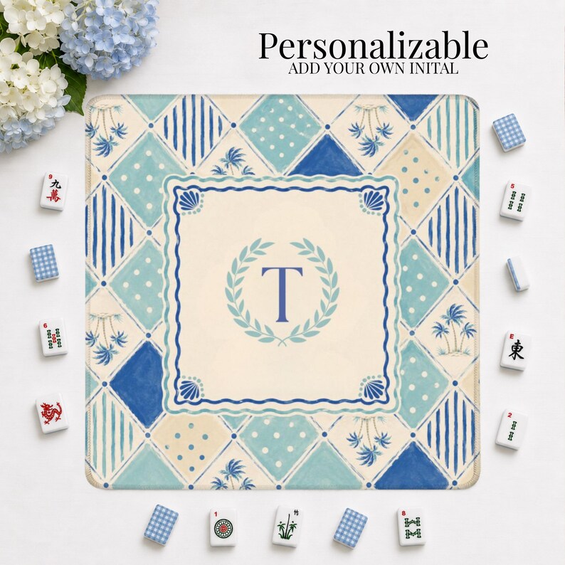 May include: A square, personalized Mahjong mat with a blue and white geometric pattern. The mat features a central design with the letter "T" inside a wreath. The text "Personalizable, Add Your Own Initial" is at the top. Mahjong tiles surround the mat.
