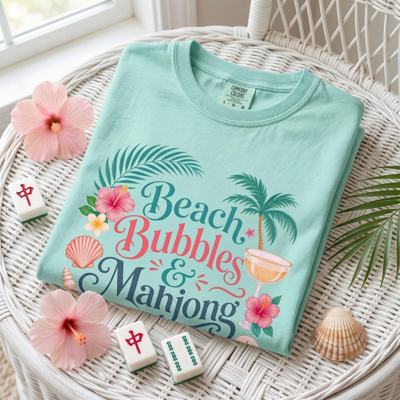 May include: A light teal t-shirt with the words "Beach Bubbles & Mahjong" in a tropical design. The design includes a palm tree, hibiscus flowers, and a cocktail glass. The shirt is folded on a white wicker chair.