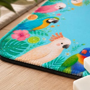 May include: A vibrant blue mousepad featuring colorful illustrations of parrots, tropical flowers, and fruit slices. The design includes a blue and gold macaw, a pink cockatoo, and a rainbow lorikeet, surrounded by green leaves and flowers.