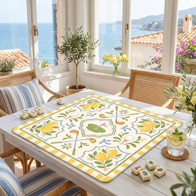 May include: A square, cream-colored table mat with a golf and lemon design. The mat features golf clubs, golf balls, and lemons, with a green and yellow border. The mat is on a white table, with a view of the ocean in the background.