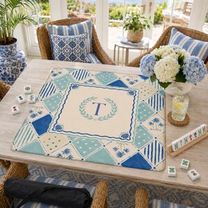 May include: A rectangular Mahjong mat with a cream-colored center featuring a blue letter "T" within a laurel wreath. The mat has a blue and white geometric pattern. The scene includes Mahjong tiles, flowers, and wicker chairs.