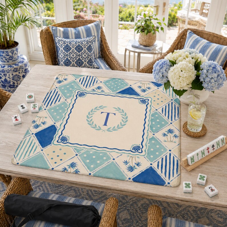 May include: A rectangular Mahjong mat with a cream-colored center featuring a blue letter "T" within a laurel wreath. The mat has a blue and white geometric pattern. The scene includes Mahjong tiles, flowers, and wicker chairs.