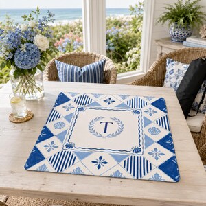 Op de afbeelding: Een vierkante placemat met een blauw en wit patroon. Het midden heeft de letter 'T' in een lauwerkrans. De placemat heeft een kustthema, met schelp- en bloemmotieven.