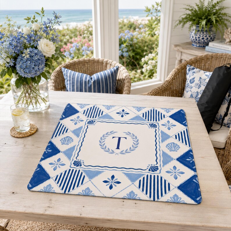 Op de afbeelding: Een vierkante placemat met een blauw en wit patroon. Het midden heeft de letter 'T' in een lauwerkrans. De placemat heeft een kustthema, met schelp- en bloemmotieven.