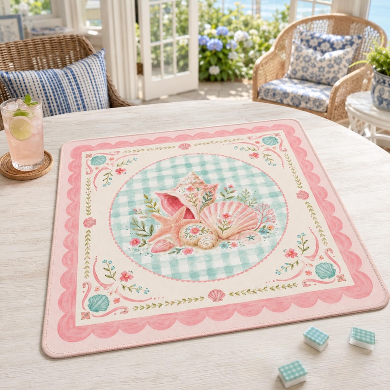 May include: A square, pink and white placemat with a scalloped edge. The center features a light blue gingham circle with seashells, starfish, and floral accents. The border has a repeating seashell and floral design.
