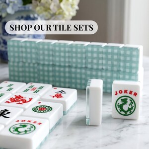 May include: A collection of white Mahjong tiles with red and green symbols and characters. Some tiles have a green dragon symbol. The text "SHOP OUR TILE SETS" is displayed on a white banner. The tiles are arranged on a white surface.