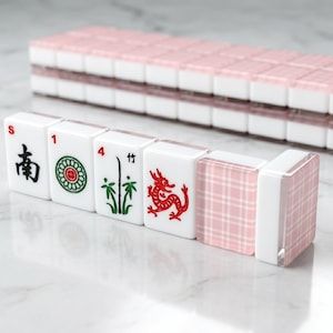 May include: A set of white Mahjong tiles featuring red, green, and black symbols and characters. The tiles are arranged in a row, with a pink and white plaid tile at the end. A stack of tiles is visible in the background.