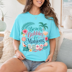 May include: A turquoise t-shirt featuring the text "Beach Bubbles & Mahjong" in a tropical design. The graphic includes palm trees, flowers, seashells, and mahjong tiles. The shirt has a relaxed fit.
