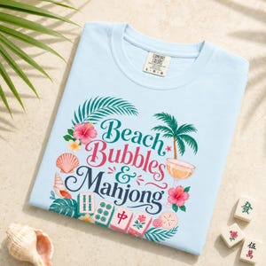 May include: Pale blue t-shirt featuring the text "Beach Bubbles & Mahjong" in a tropical design. The graphic includes palm trees, flowers, seashells, and mahjong tiles. The shirt is a Comfort Colors brand.