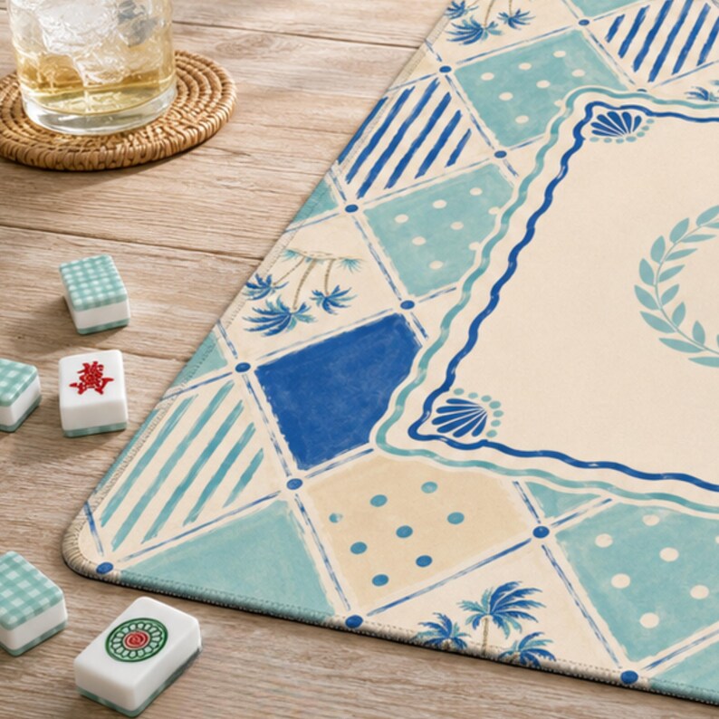 May include: A blue and white patterned Mahjong mat with a decorative border and palm tree accents. Several Mahjong tiles are scattered around the mat, with a glass of water and a coaster in the background. The mat has a coastal theme.