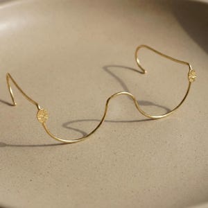 Puede incluir: Una delicada diadema de alambre dorado con un diseño ondulado. La diadema presenta dos pequeños elementos decorativos circulares en cada extremo. El metal tiene un acabado pulido, que refleja la luz. La pieza está diseñada para ser usada en la cabeza.