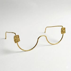 Könnte beinhalten: Eine goldfarbene Drahtgestell-Brille mit einem einzigartigen Design. Der Rahmen hat einen wellenförmigen Steg und rechteckige dekorative Elemente an den Bügeln. Die Bügel haben ein kleines, quadratisches Design. Die Gesamtästhetik ist minimalistisch und elegant.