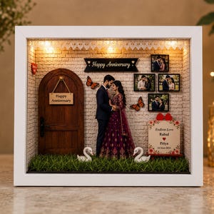 May include: A white shadow box frame containing a miniature scene with a wooden door, a couple figurine, two white swan figures, and artificial grass. The wall features a Happy Anniversary sign, three framed photos, and a sign reading Endless Love Rahul Priya.