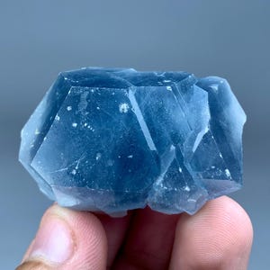 May include: A raw, deep blue fluorite crystal cluster featuring multiple geometric, translucent facets. The mineral specimen displays varying shades of blue with internal inclusions and sharp, angular edges throughout its crystalline structure.