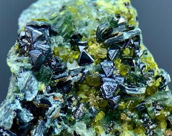 Könnte beinhalten: Ein Mineralexemplar mit einer Ansammlung dunkler, metallischer, dreieckiger Kristalle, die in eine Matrix aus durchscheinenden, limonengrünen und olivgrünen Kristallstrukturen eingebettet sind. Die Oberfläche zeigt scharfe, reflektierende dunkle Facetten.