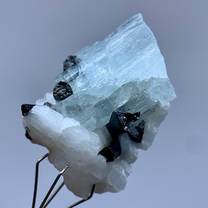 May include: A close-up of a mineral specimen featuring a large, light blue crystal formation with black accents. The crystal is supported by a white, crystalline base. The specimen is held by a metal stand against a soft gray background.