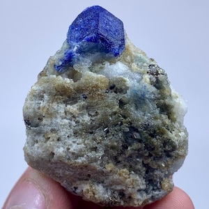 May include: A raw, natural lapis lazuli specimen. The deep blue crystal is faceted and sits atop a matrix of beige, gray, and white minerals. Small flecks of pyrite add a metallic shimmer to the stone. The overall appearance is rough and organic.