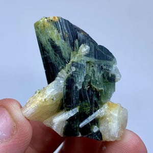 May include: A close-up of a mineral specimen featuring a cluster of dark green and black crystals with a striated texture. The crystals are accented with lighter green and white crystalline formations, and a touch of yellow.